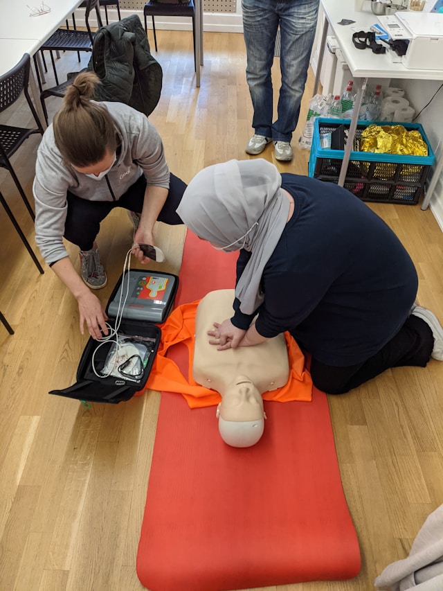 A woman doing chest compressions on a CPR training doll while another gets a defibrillator ready. 